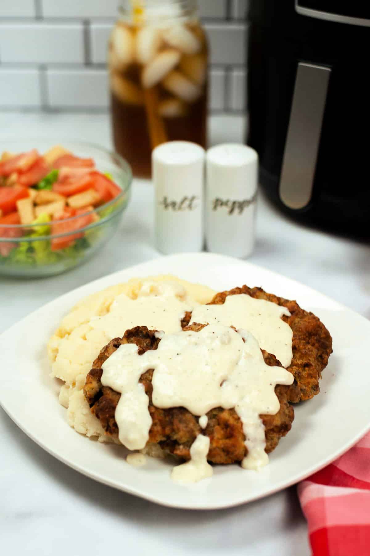 chicken fried steak with gravy on a white plate with a bowl of salad, salt and pepper shakers, a pitcher of tea, and an air fryer in the background