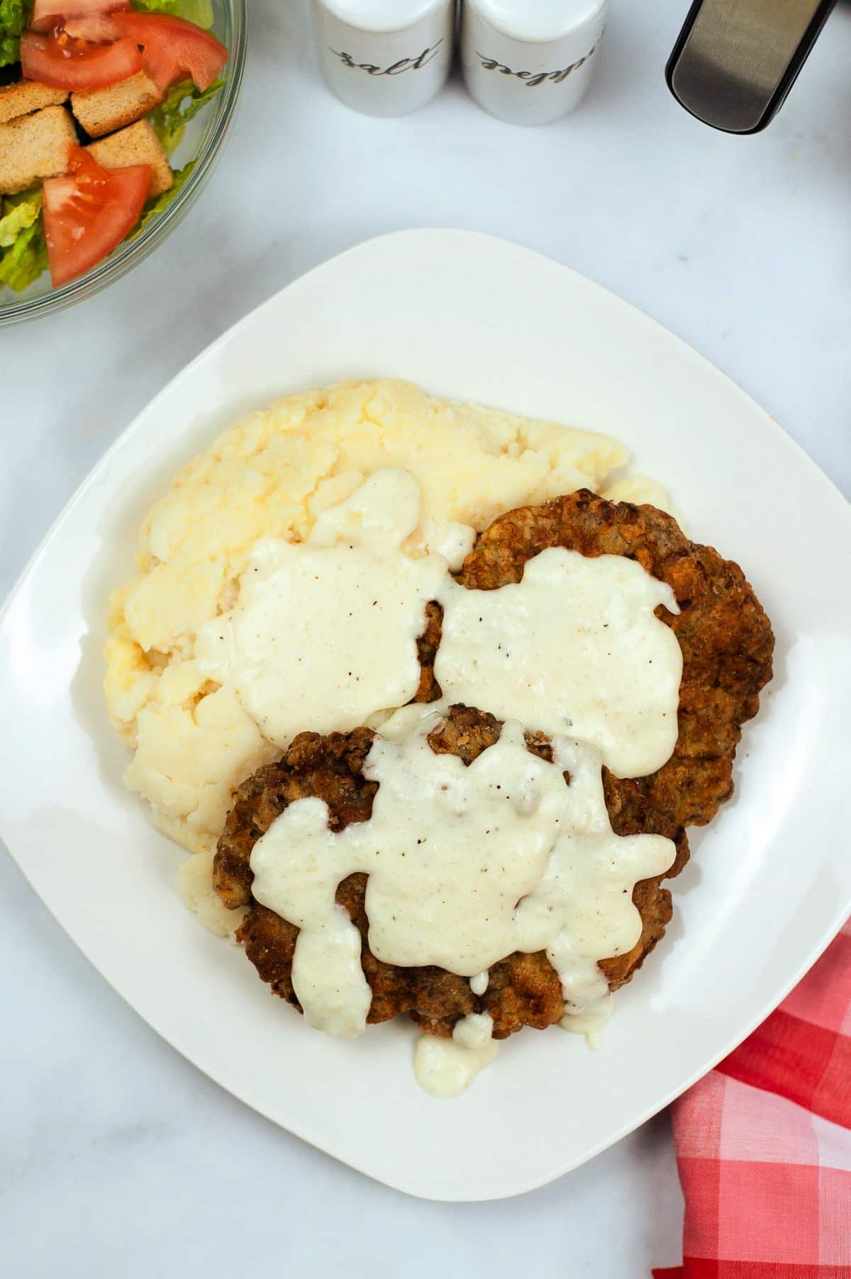 overhead view of chicken fried steak with gravy on a white plate with a bowl of salad, salt and pepper shakers, and an air fryer in the background