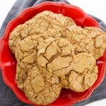 A red plate is filled with a stack of large, cracked peanut butter cookies, evoking the golden hue of classic carrot cookies, all set against a gray cloth backdrop.