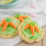 Carrot Sugar Cookies adorned with green coconut shreds and cute carrot-shaped candies rest on a white cloth, blending flavor and whimsy.