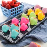 A tray of Easter chocolate-covered strawberries dipped in colorful pastel coatings. A basket of plain strawberries sits in the background on a blue cloth, adding a touch of festive elegance.