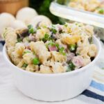 A bowl of pasta salad with ham tetrazzini, featuring peas, mushrooms, and parsley, topped with grated cheese. A striped cloth and onions are partially visible in the background.