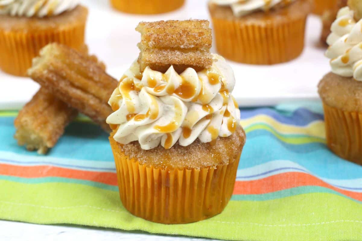 Cinnamon Cupcakes on a colorful tablecloth, garnished with caramel sauce and a piece of churro.
