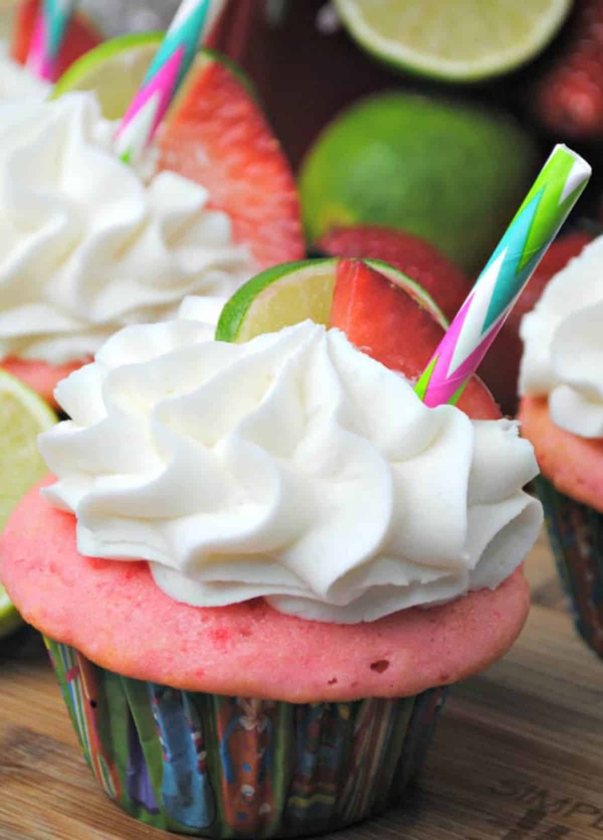 Strawberry Margarita Cupcakes topped with lime and strawberry and colorful straw on a wooden board.