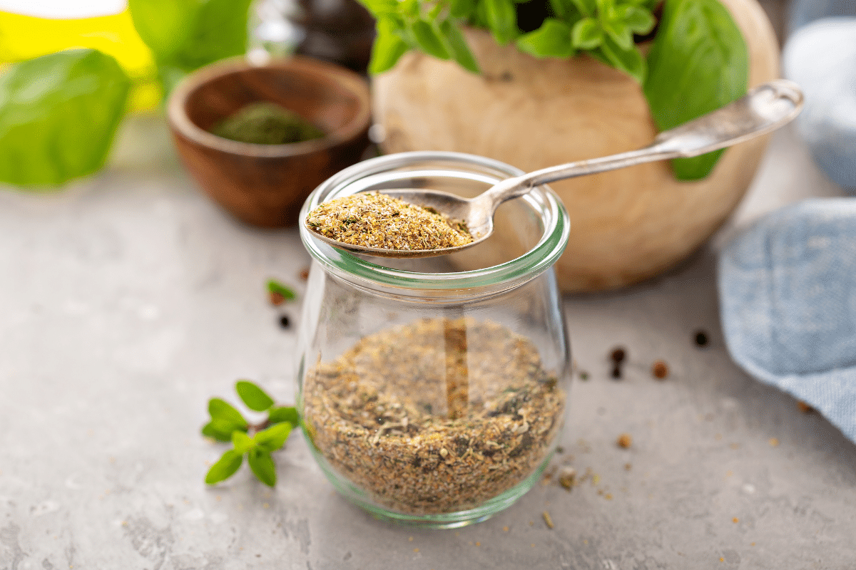 Greek seasoning mix in a glass jar, with spoon on top with seasoning mix.
