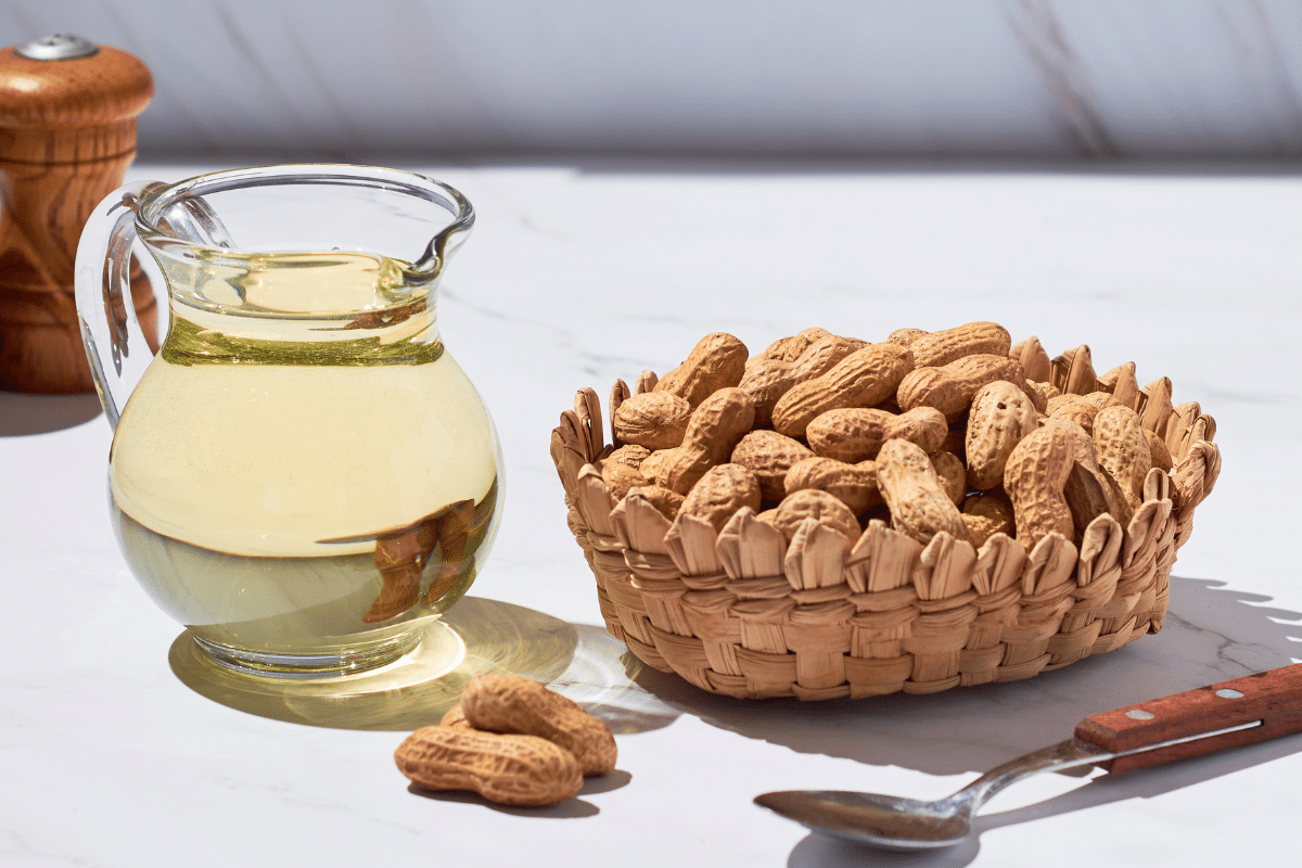 A glass jar of peanut oil with a basket full of peanuts and a spoon on the side.