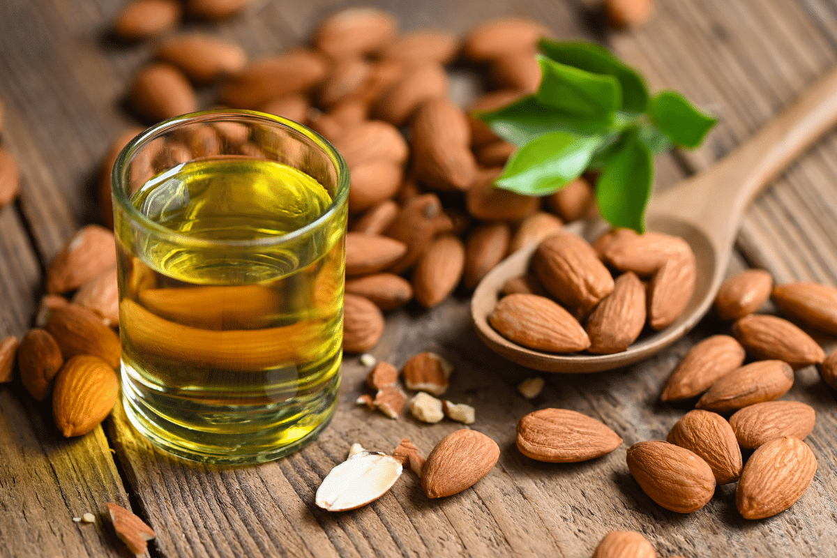 Almond oil in a small glass, almonds on a wooden spoon on the side.