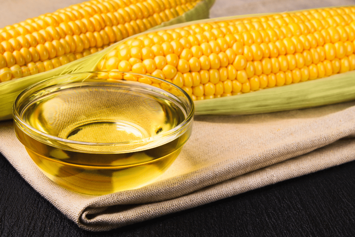 Corn oil in a small bowl sits on a rustic cloth, with corn on the side.