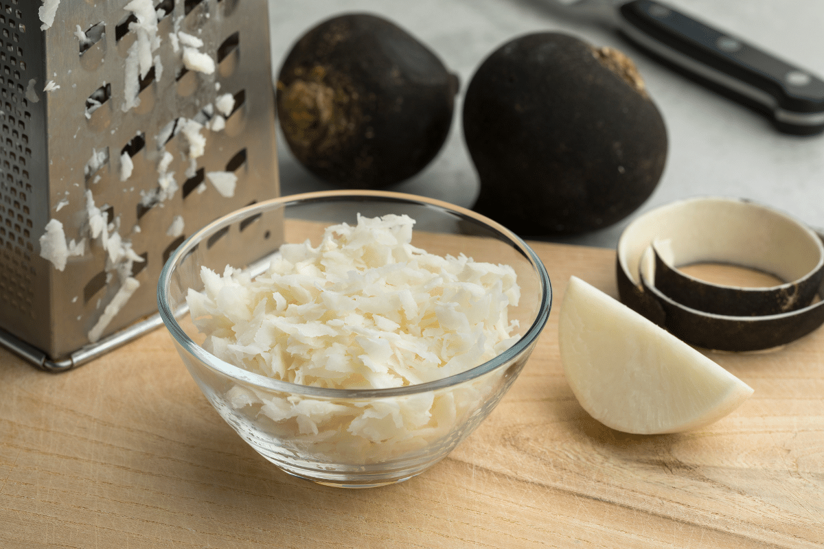 Grated black radish in a small bowl with grater, knife and sliced black radish on the side.