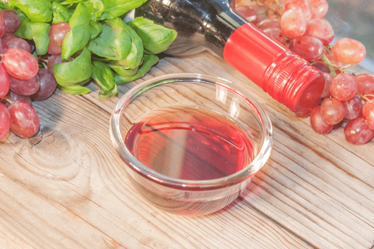 A bowl of red wine vinegar on a wooden table with grapes, wine and basil on the side.