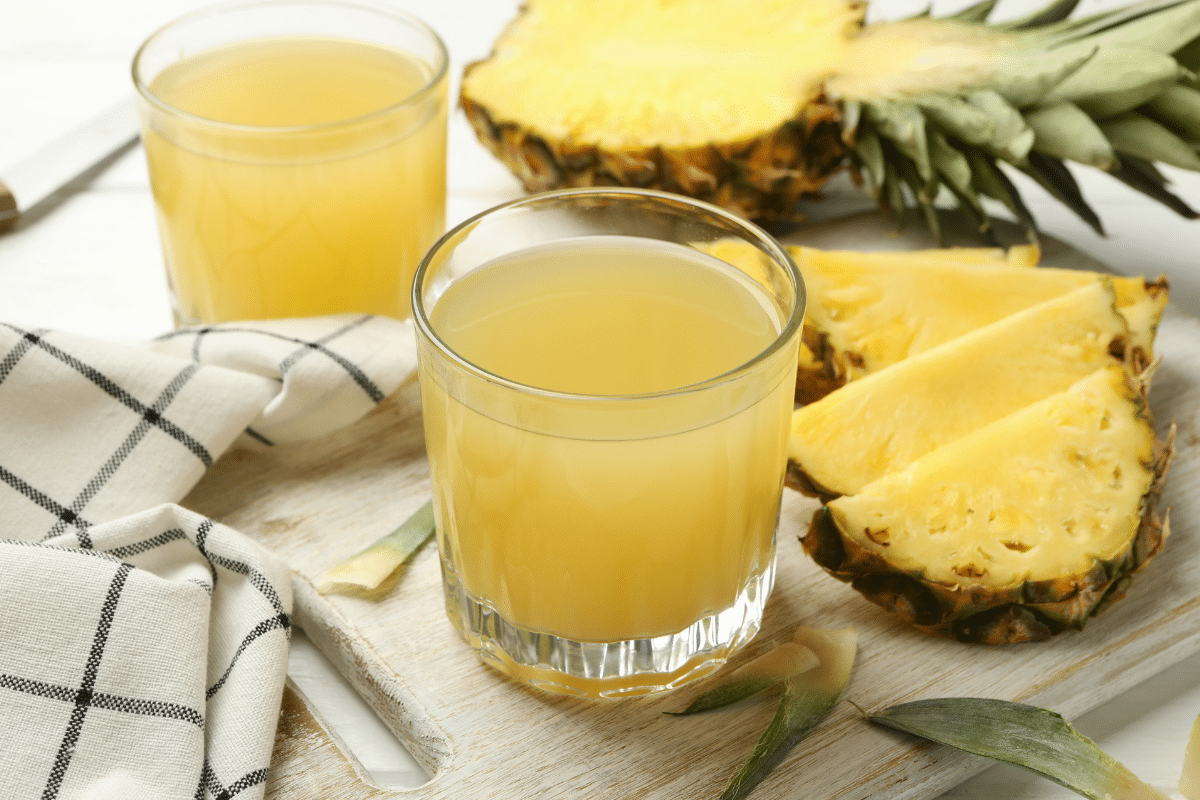 Two cups of pineapple juice on a wooden board, with fresh pineapples on the side.