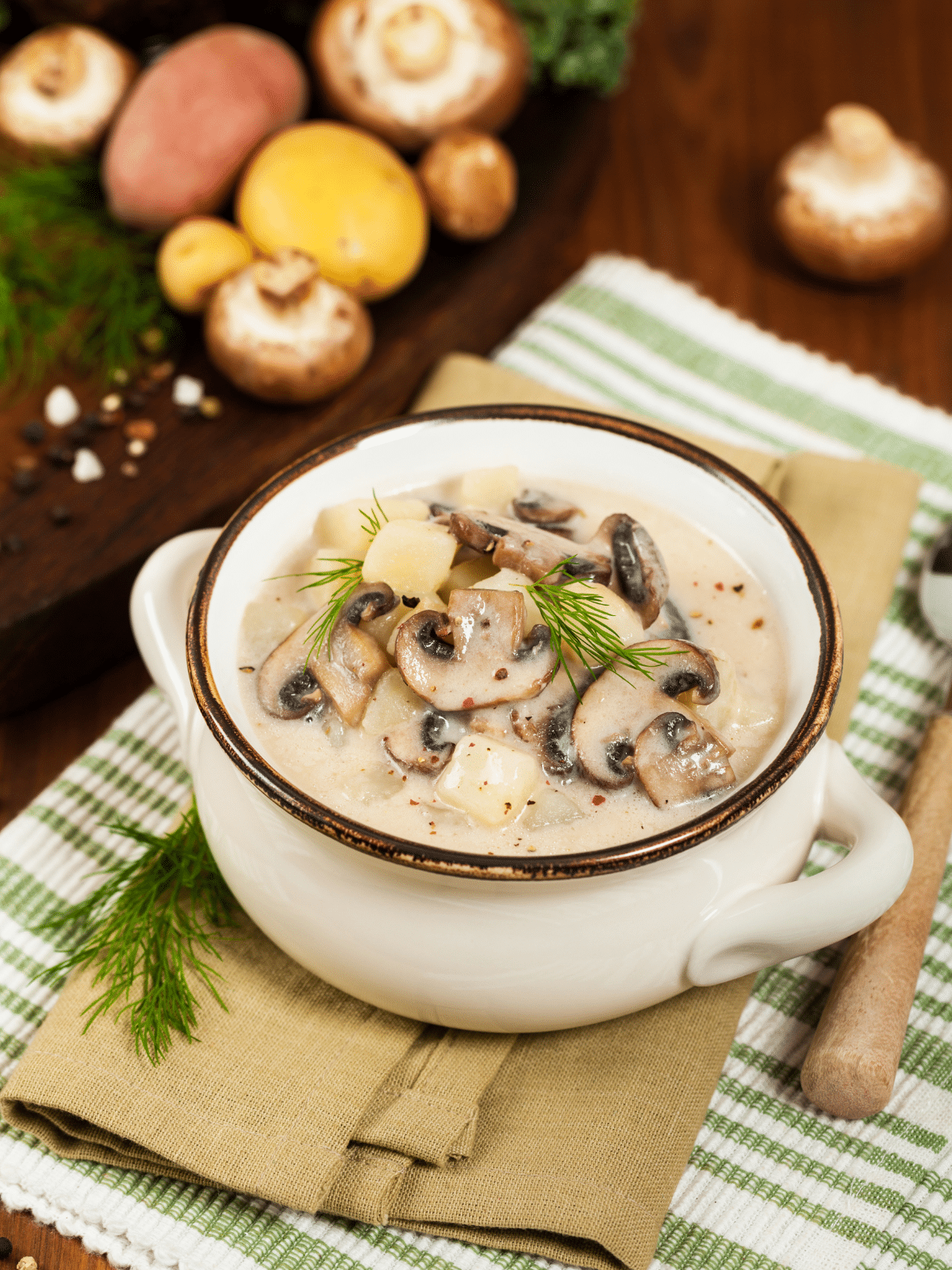 A bowl of cream of mushroom soup on a tablecloth with mushrooms and spoon on the side.