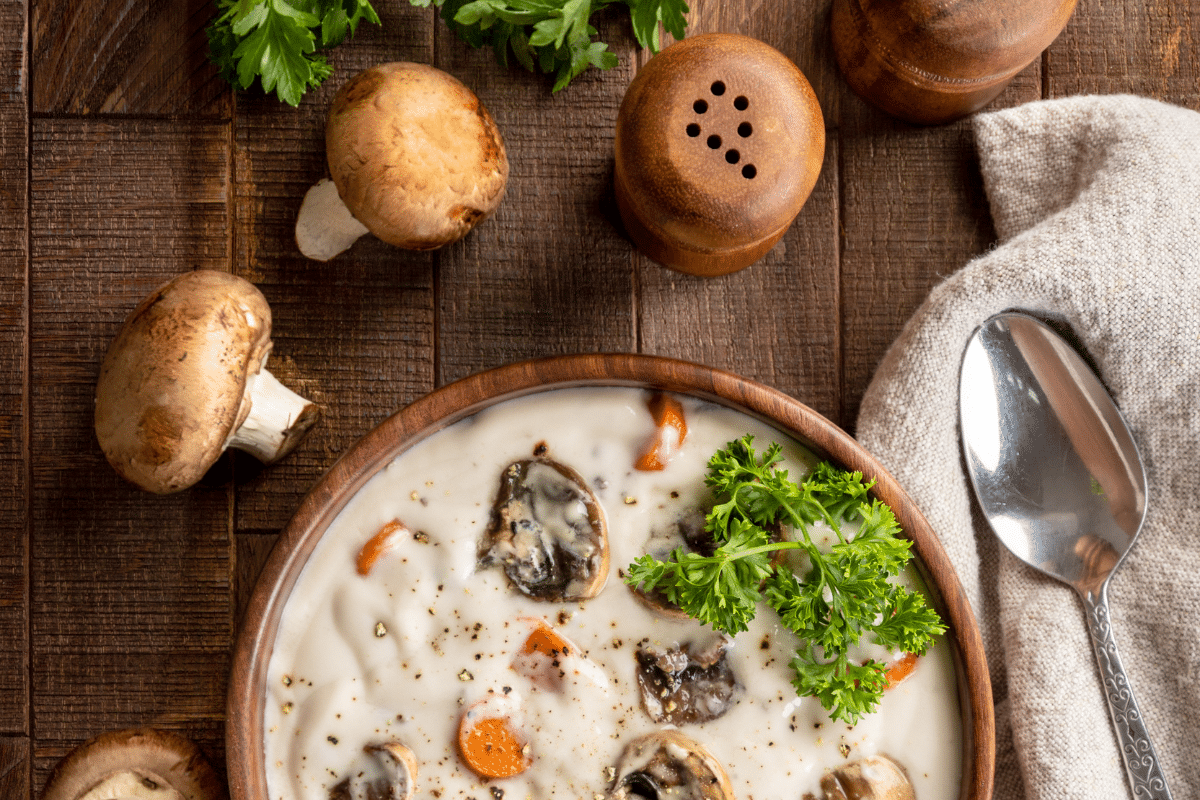 A bowl of cream of mushroom soup on a wooden table with Mushrooms and spoon on the side.