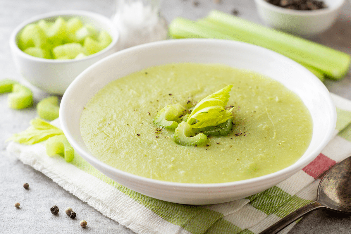 A bowl of cream of celery soup on a tablecloth with celery and spoon on the side.