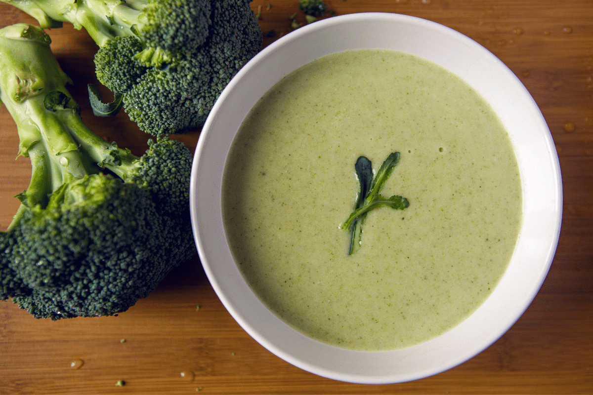 A bowl of cream of broccoli soup on a wooden board with broccoli on the side.
