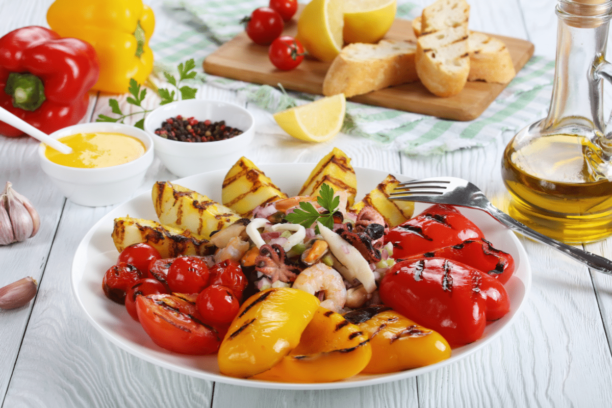 Seafood and grilled vegetables on a white plate next to a jar of oil and other ingredients.