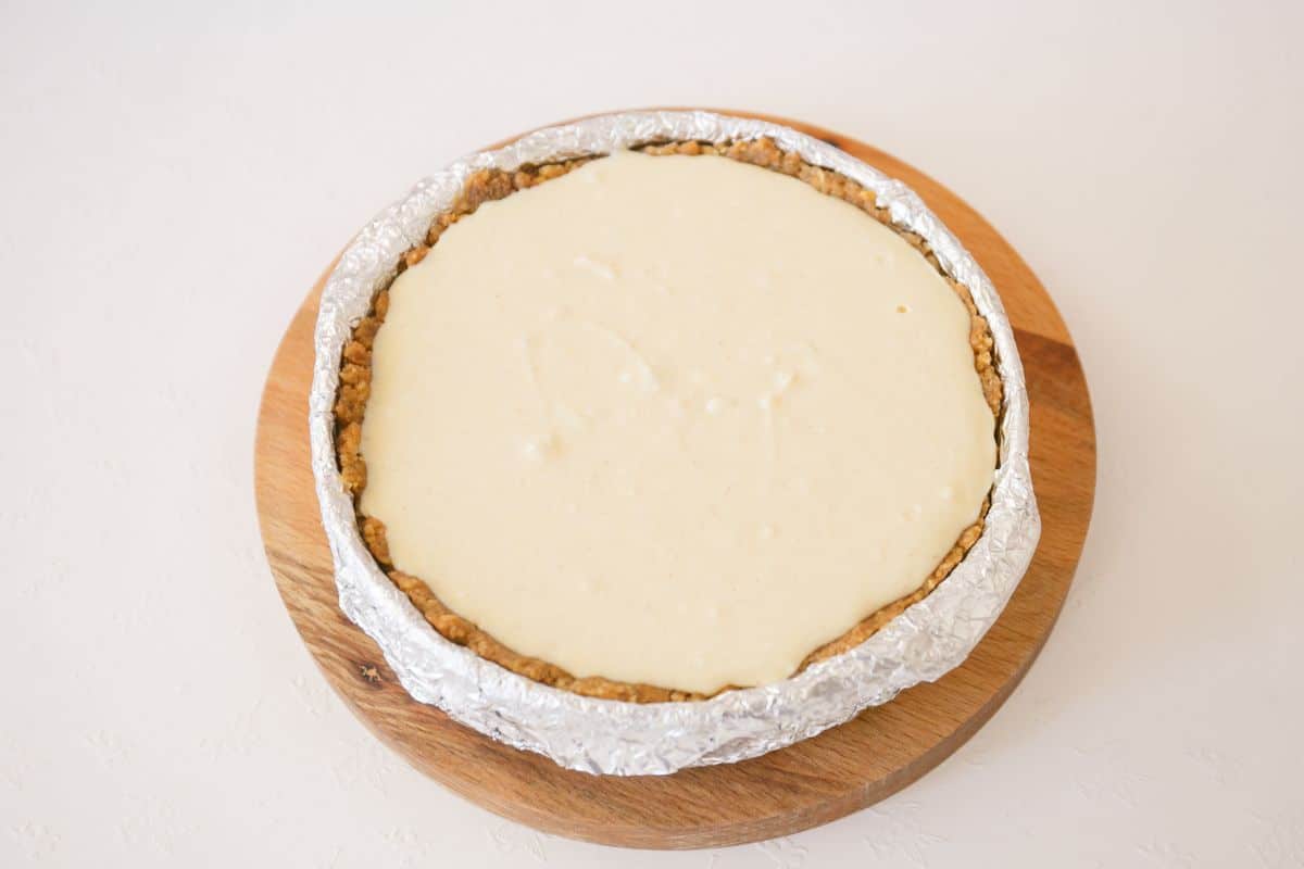 Cheesecake crust and filling in a round pan with foil on a wooden board.