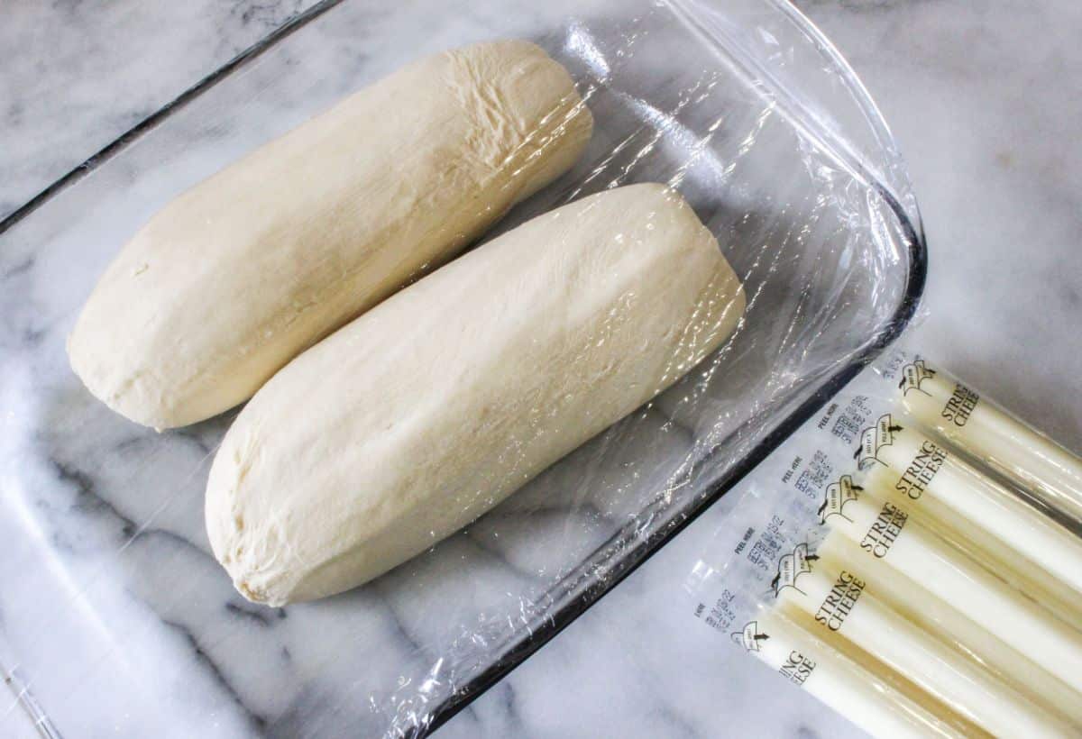 Two frozen bread loaves in a baking dish covered with plastic wrap, next to Mozzarella cheese sticks.