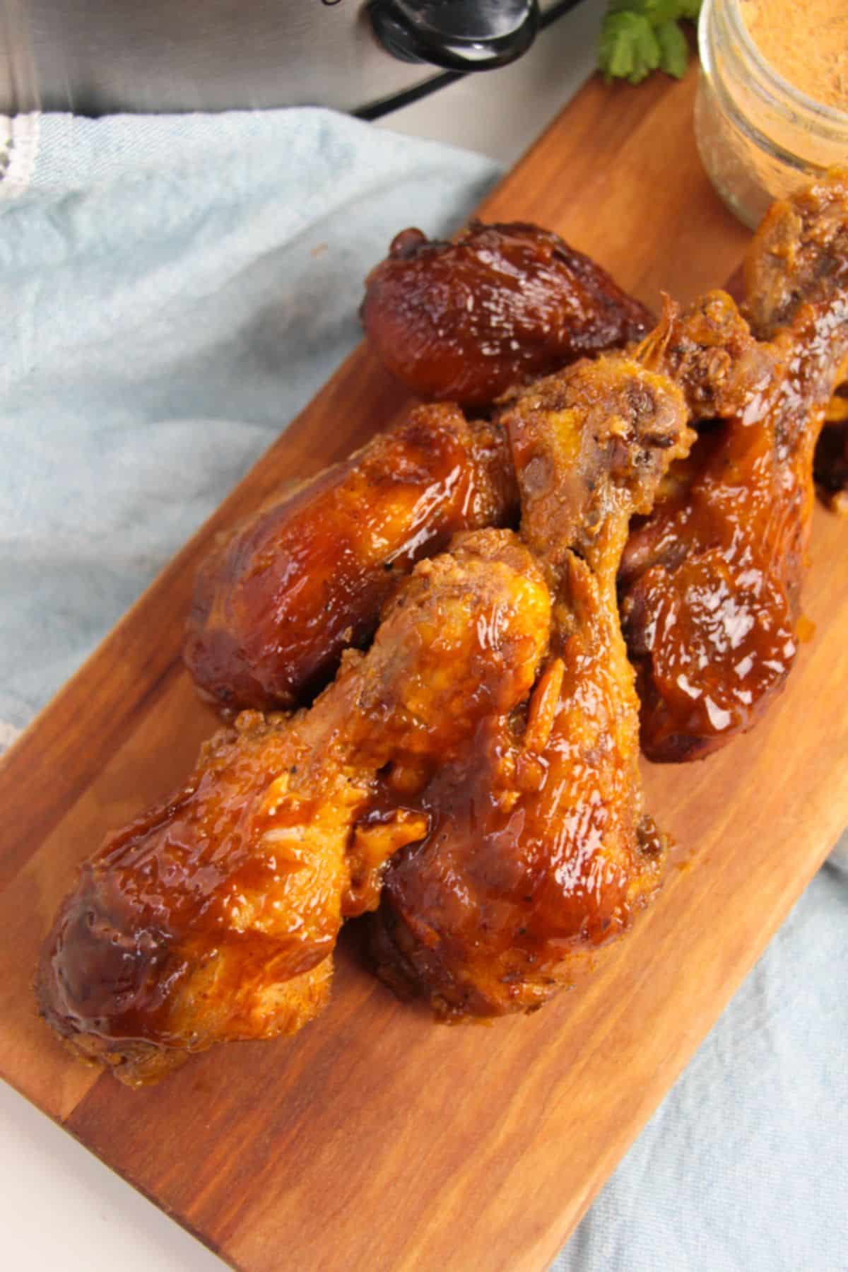 Slow Cooker BBQ Chicken Legs on a wooden board with crockpot and seasoning on the side.