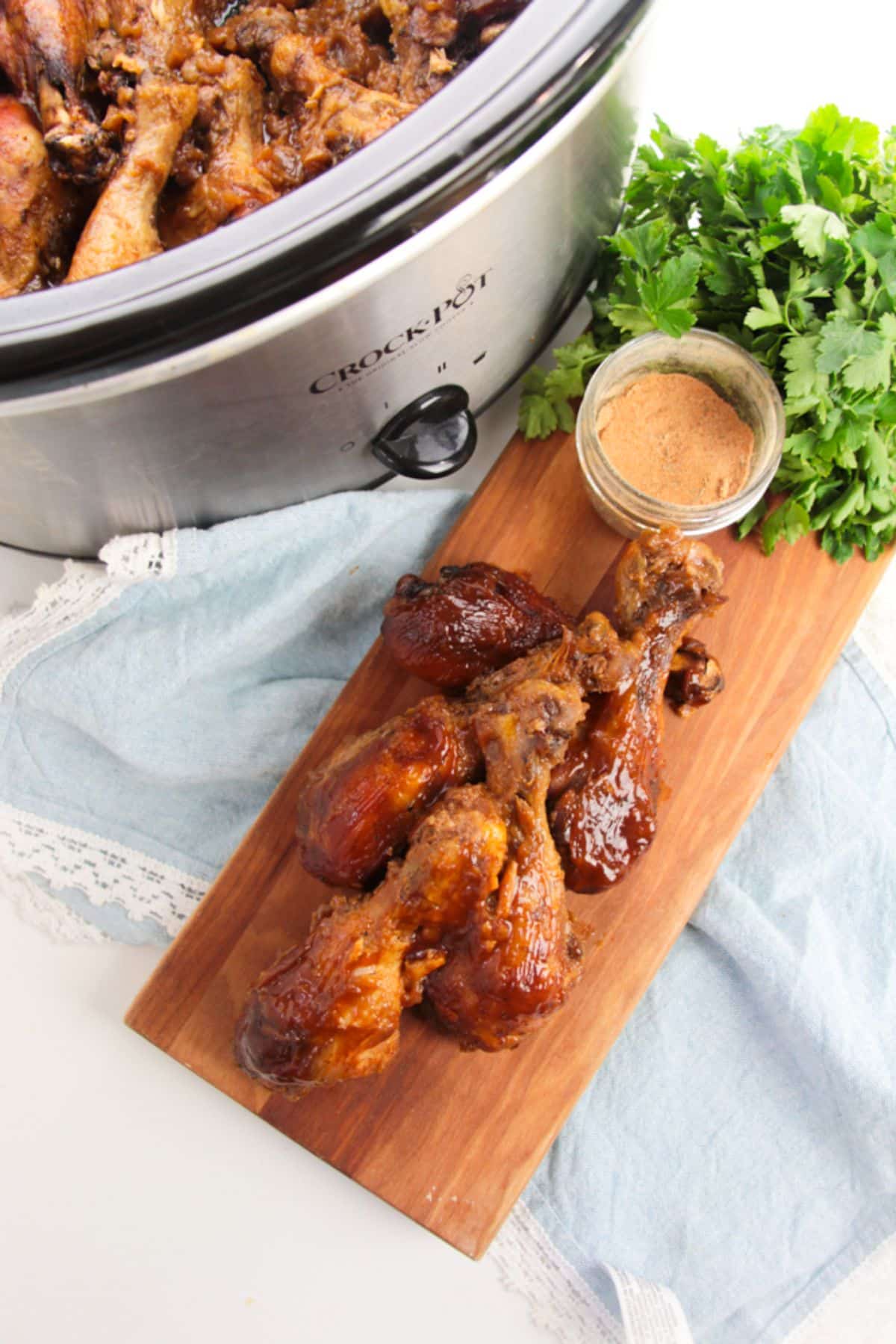 A slow cooker with BBQ chicken legs next to more chicken legs on a cutting board.
