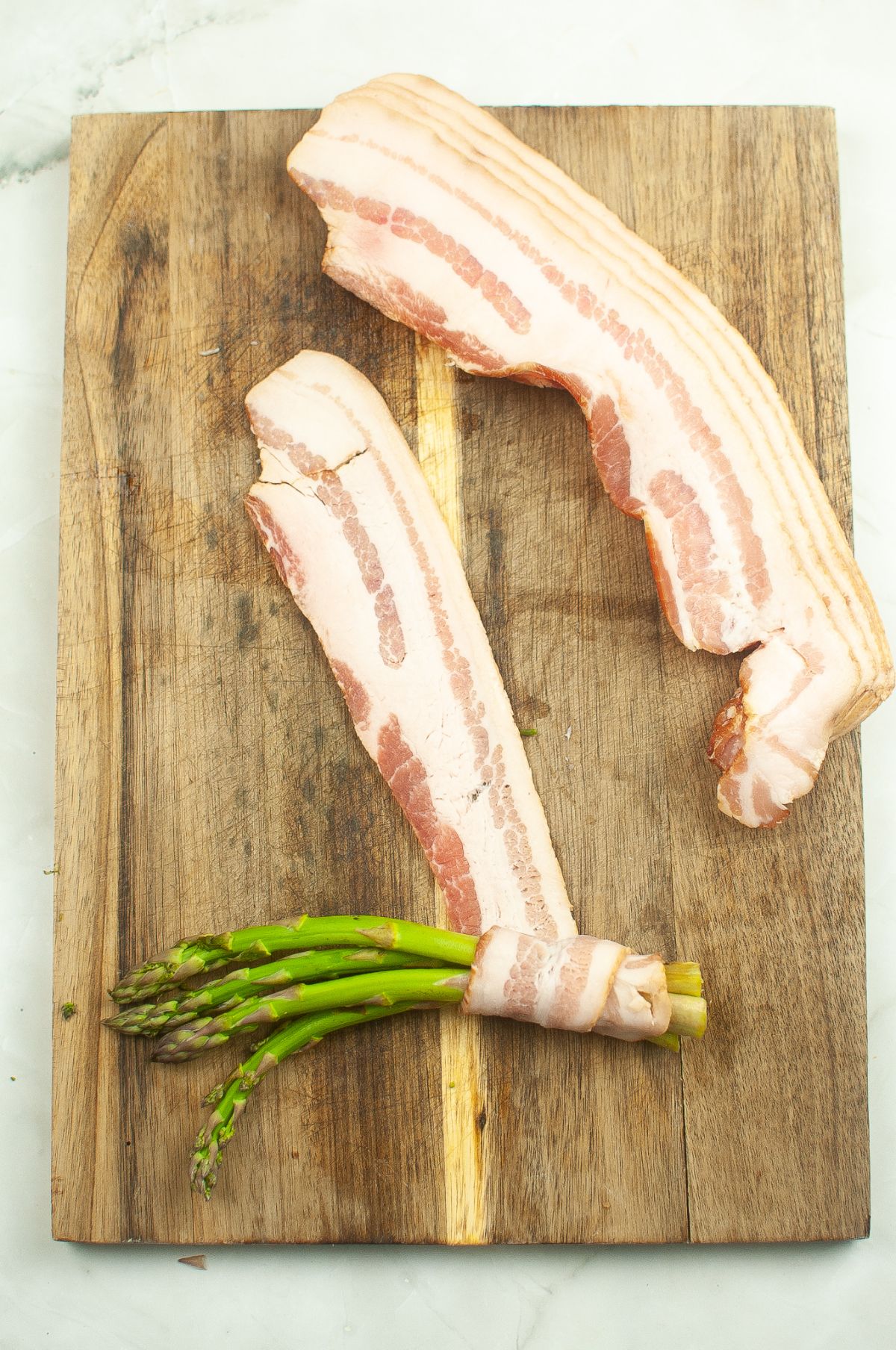 Sliced bacon being used to wrap up a bunch of asparagus on a wooden cutting board.