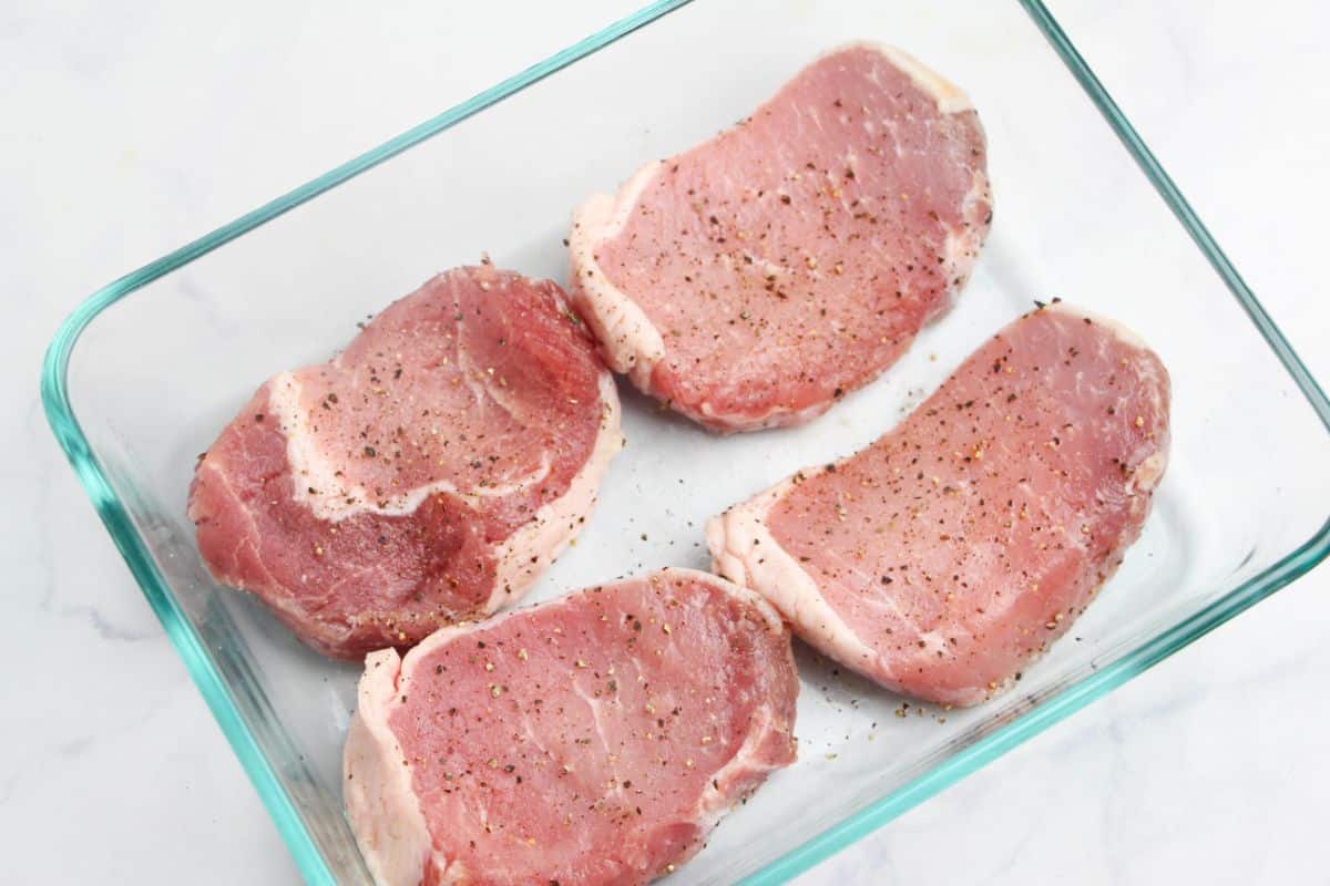 Raw pork chops seasoned with salt and pepper in a glass dish on a white countertop.