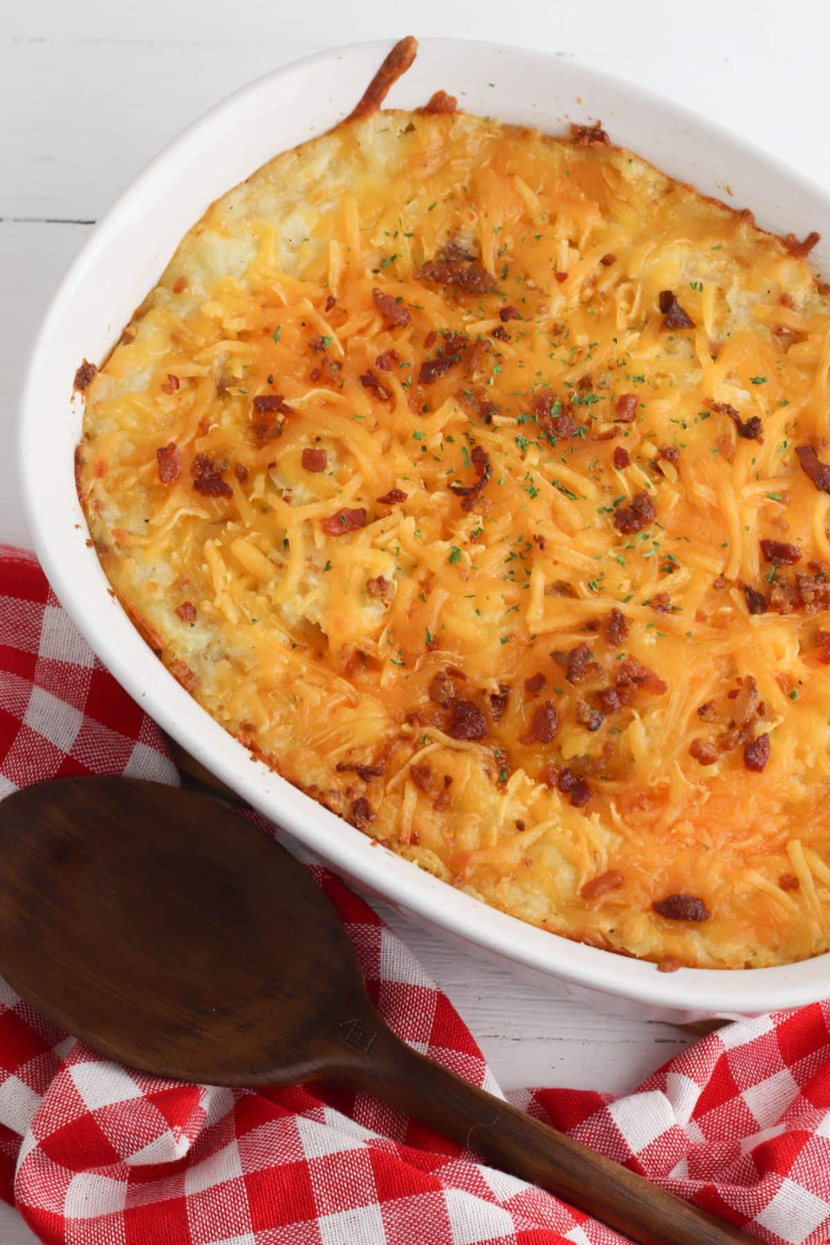 Twice Baked Loaded Potato Casserole in a baking dish. Wooden spoon with red and white checkered tablecloth on the side.