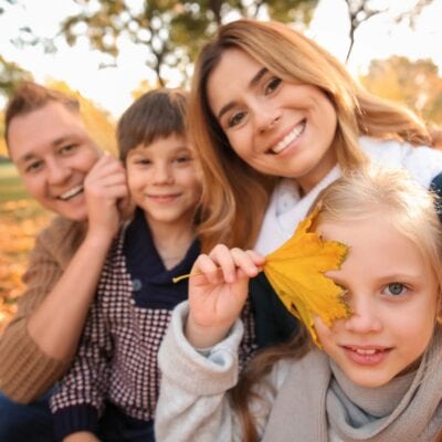 A family of four sits outdoors in autumn, smiling at the camera. The young girl in front holds a yellow maple leaf over one eye—making memories for their fall bucket list.