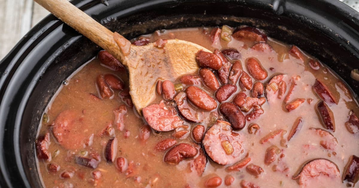 A crock pot full of beans and sausage with a wooden spoon.