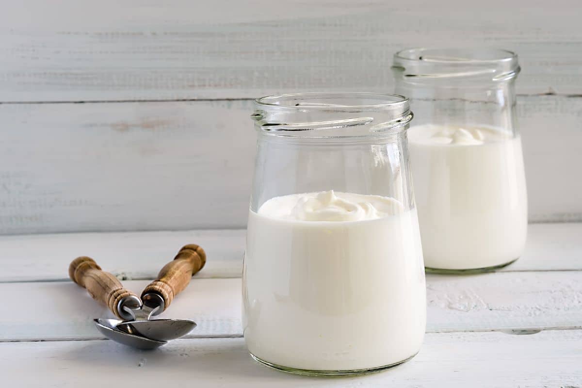 Two jars of plain yogurt with a spoons on a wooden table.