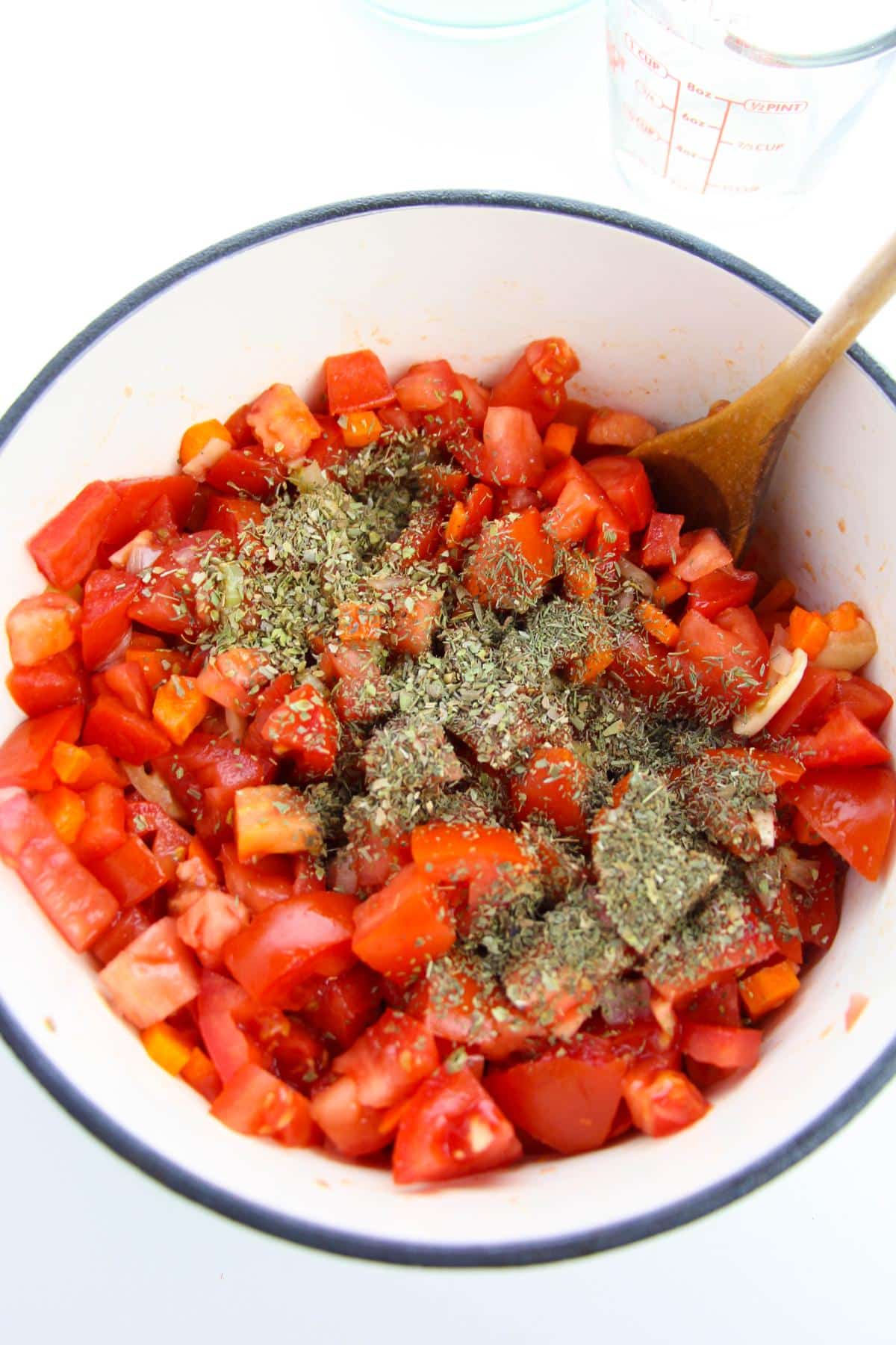 A white bowl with tomatoes and Italian seasoning in it, and a wooden spoon on the side.