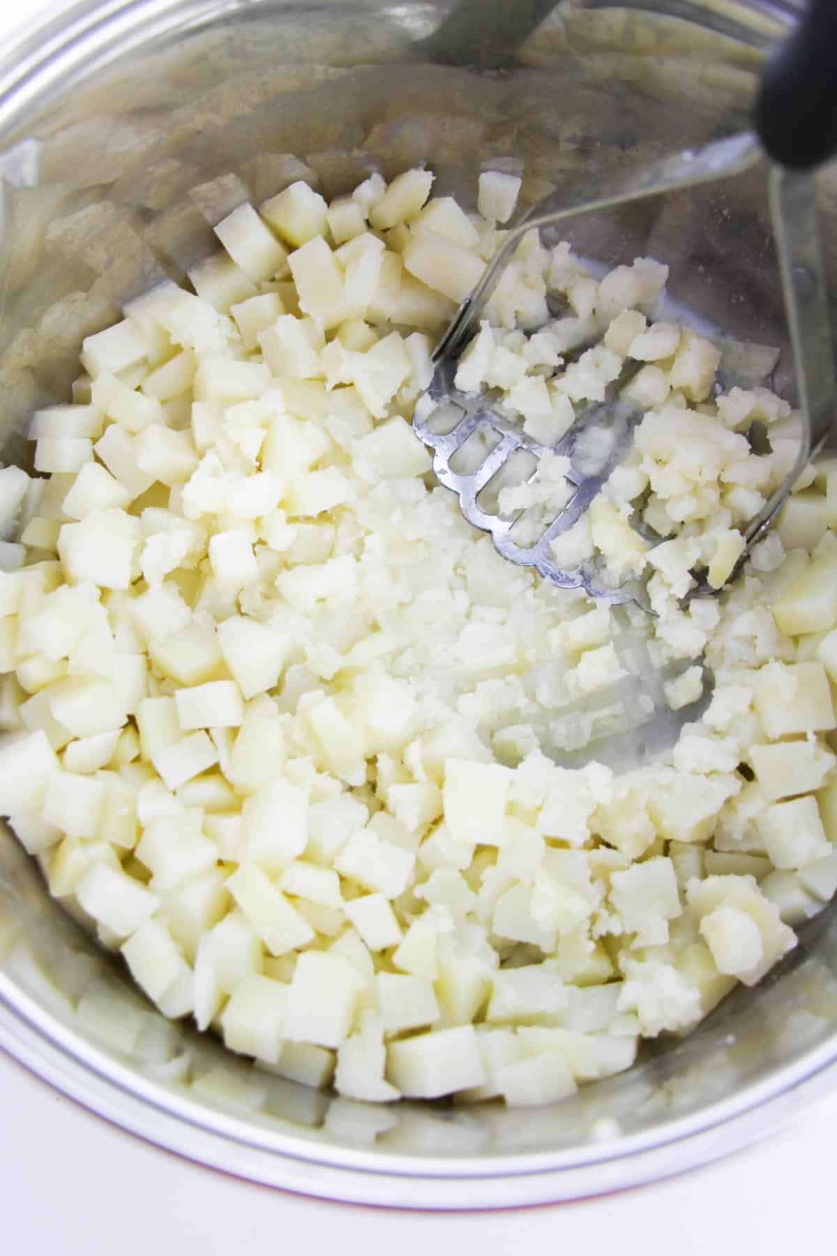 Cooked potatoes are being mashed using potato masher in a mixing bowl.