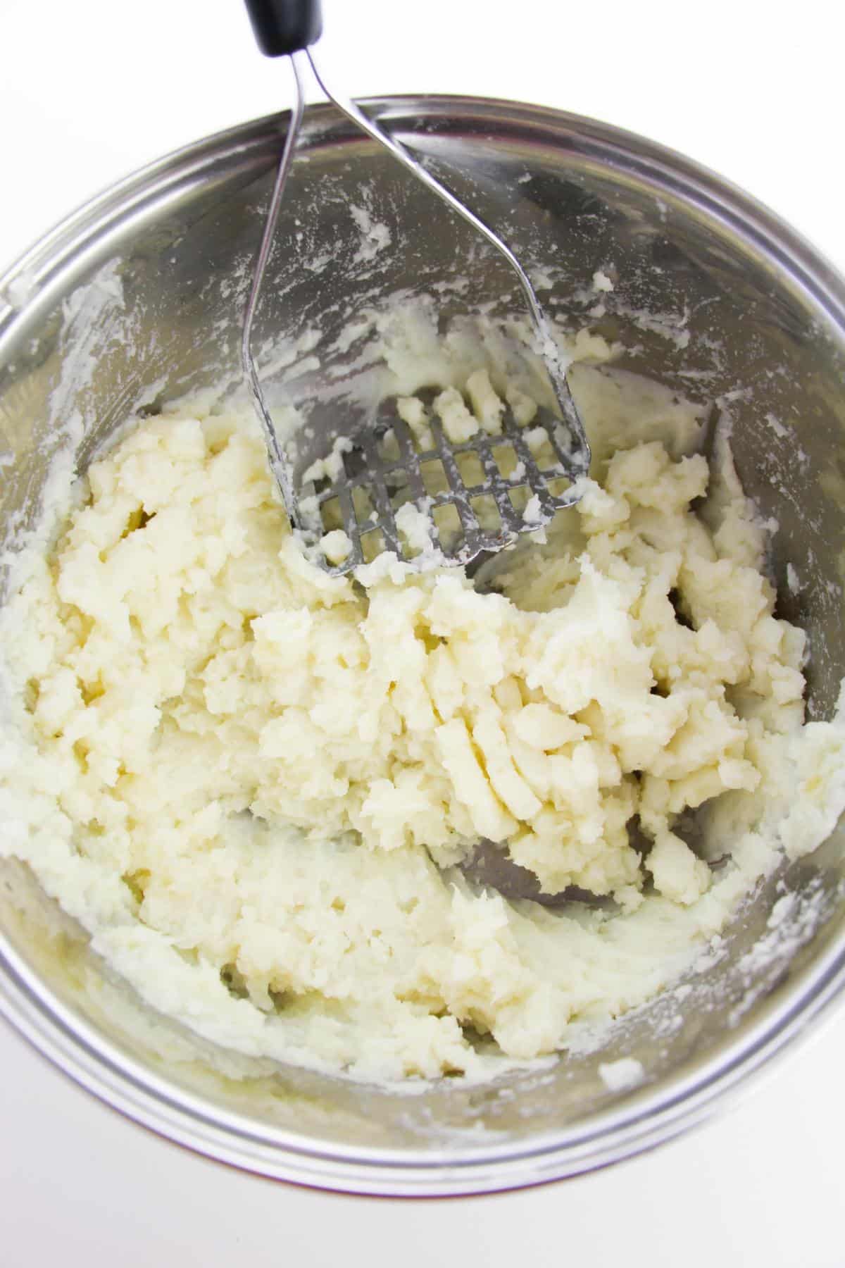 Cooked potatoes are being mashed using potato masher in a mixing bowl.