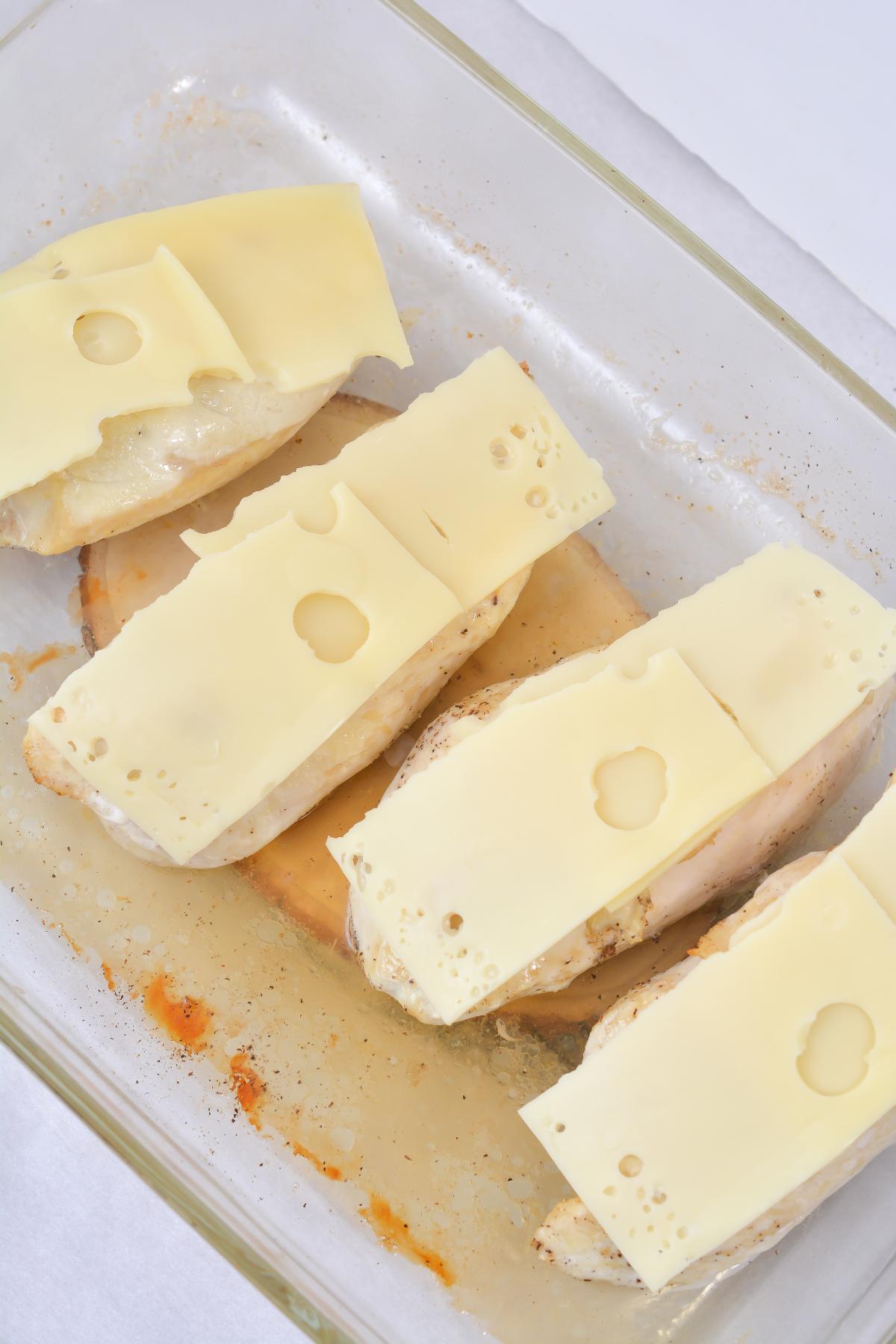 Four pieces of chicken breast in a baking dish topped with Swiss cheese.