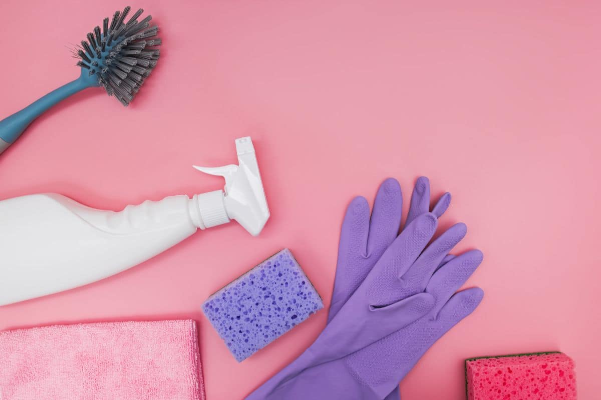 Purple cleaning gloves, spray bottle, cloth, soft brush and sponges on a pink background.