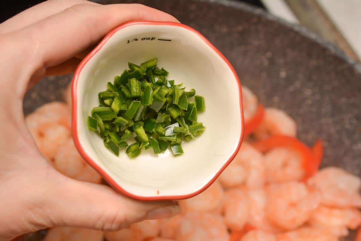 A person is holding a bowl with  serrano peppers in it, above a pan of shrimp.