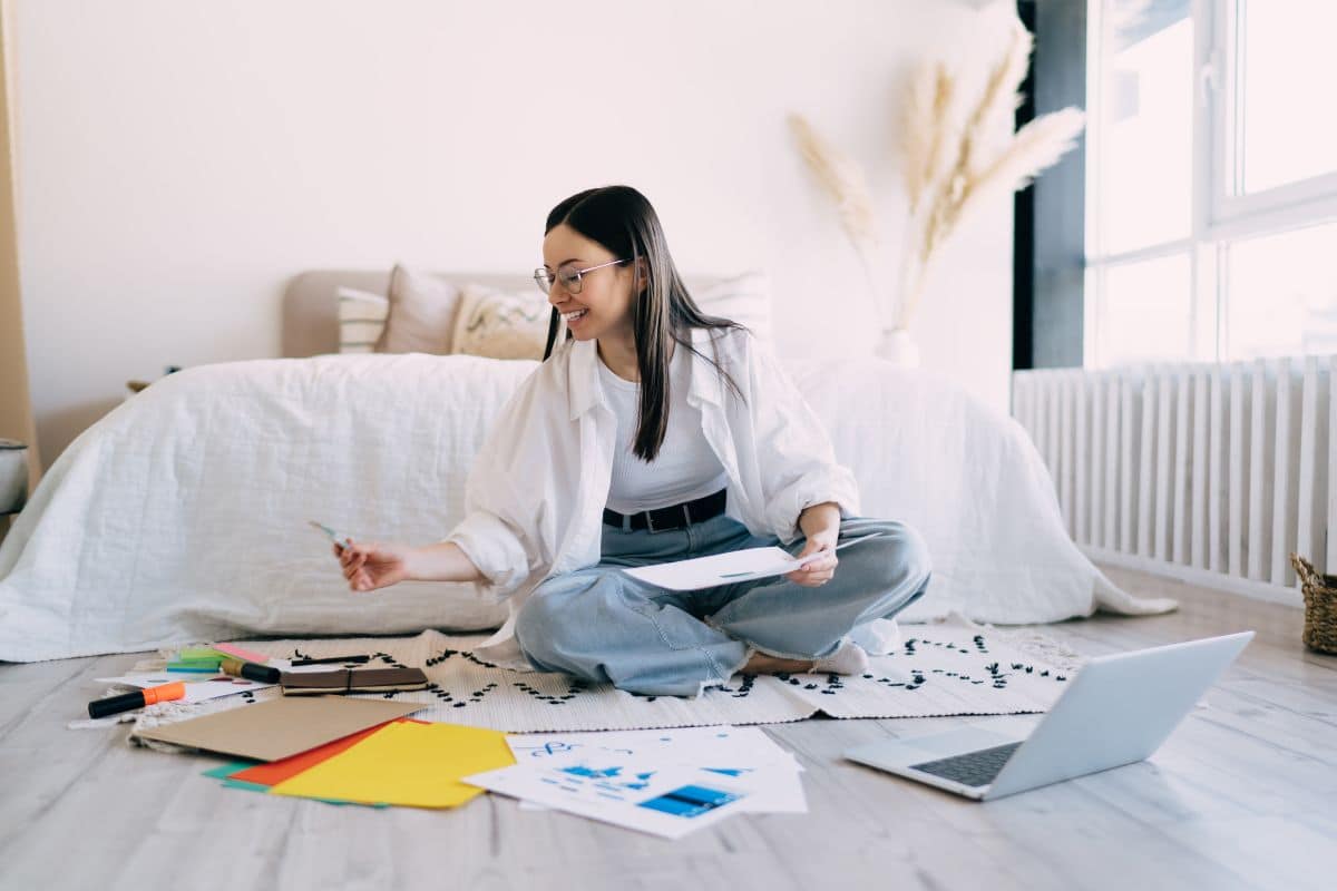 A woman sitting on the floor of her bedroom with a laptop and vision board.