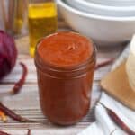 A jar of homemade chile enchilada sauce on a wooden table surrounded by ingredients.