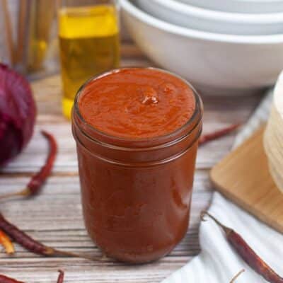 A jar of homemade chile enchilada sauce on a wooden table surrounded by ingredients.