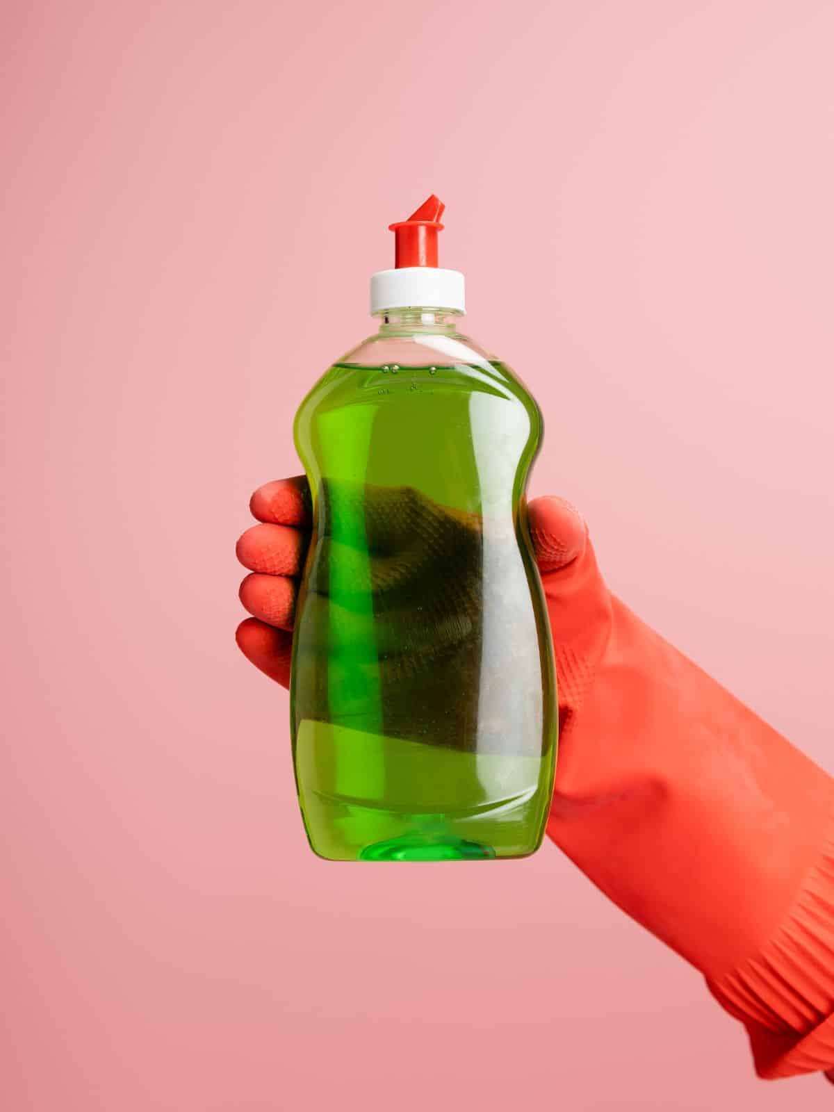 A hand holding a bottle of green dishwashing liquid on a pink background.