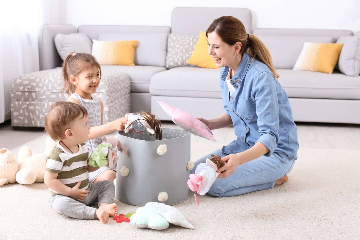 A mother and two children tidying up stuffed animals in a living room.