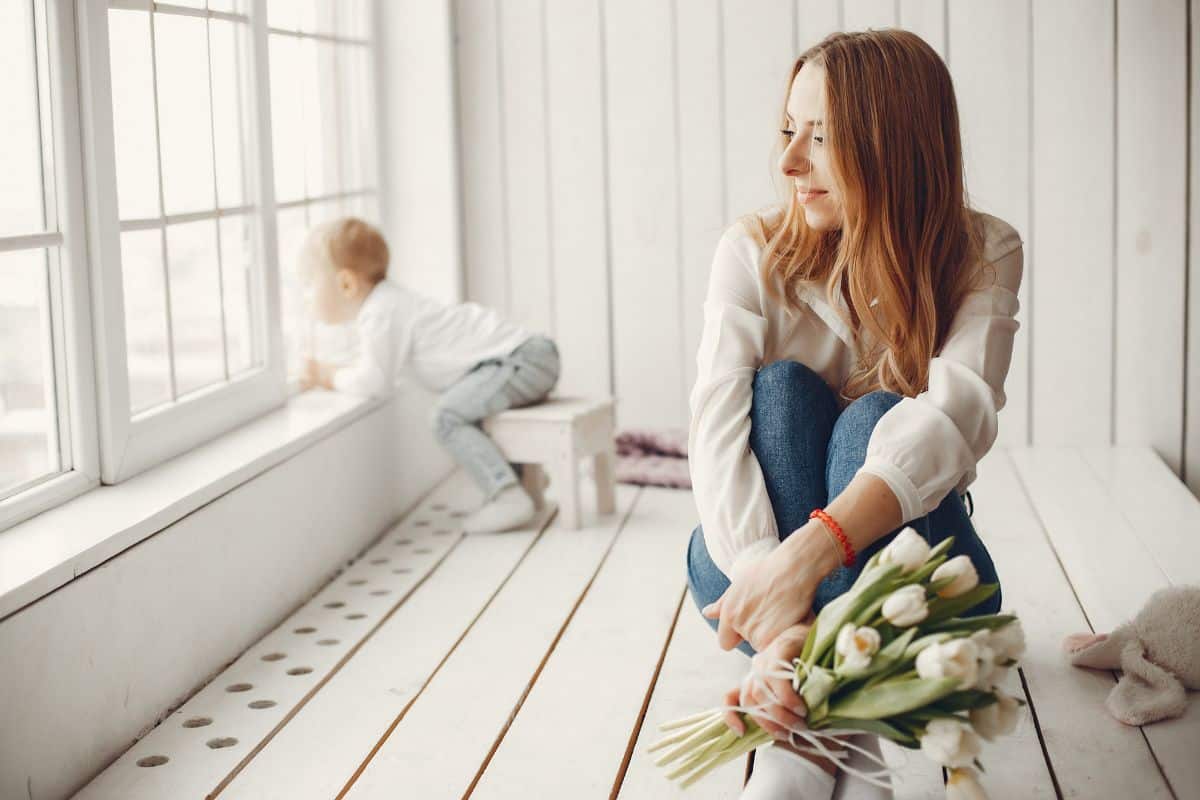 A woman sits on the floor next to her son and window with a bouquet of tulips.