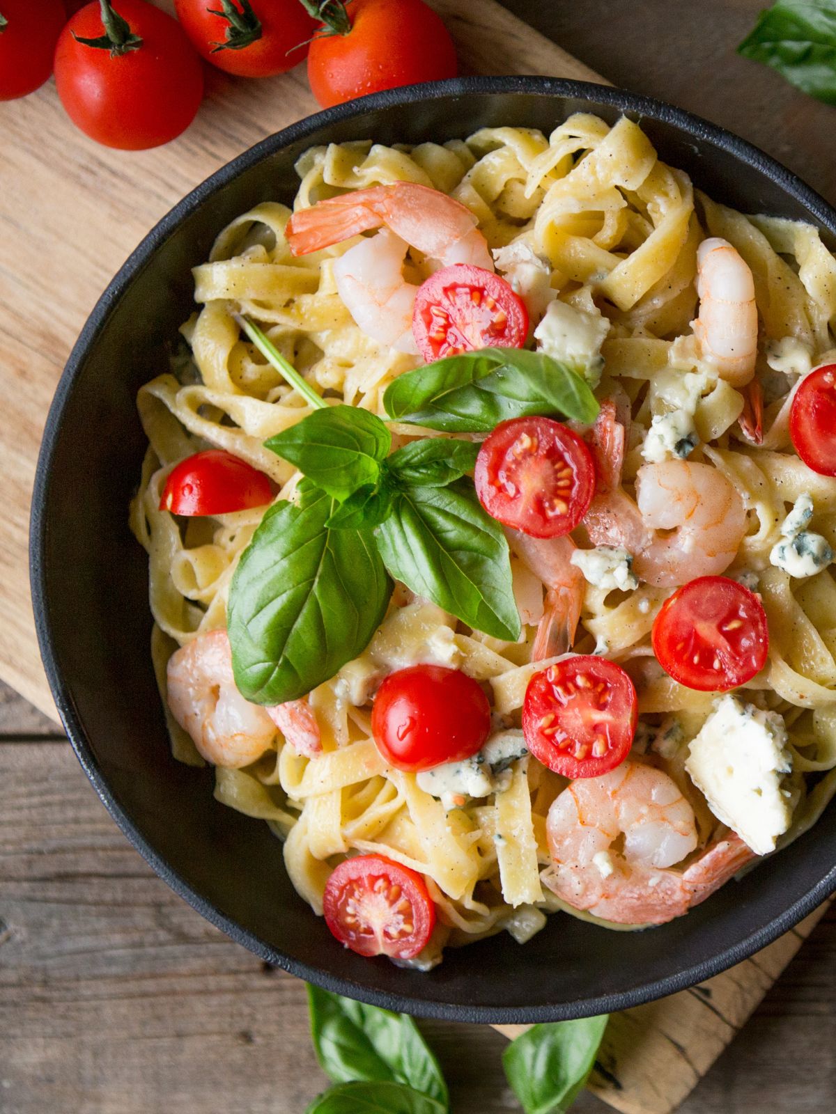 Pasta with shrimp and tomatoes in a skillet on a wooden table, garnished with fresh basil.
