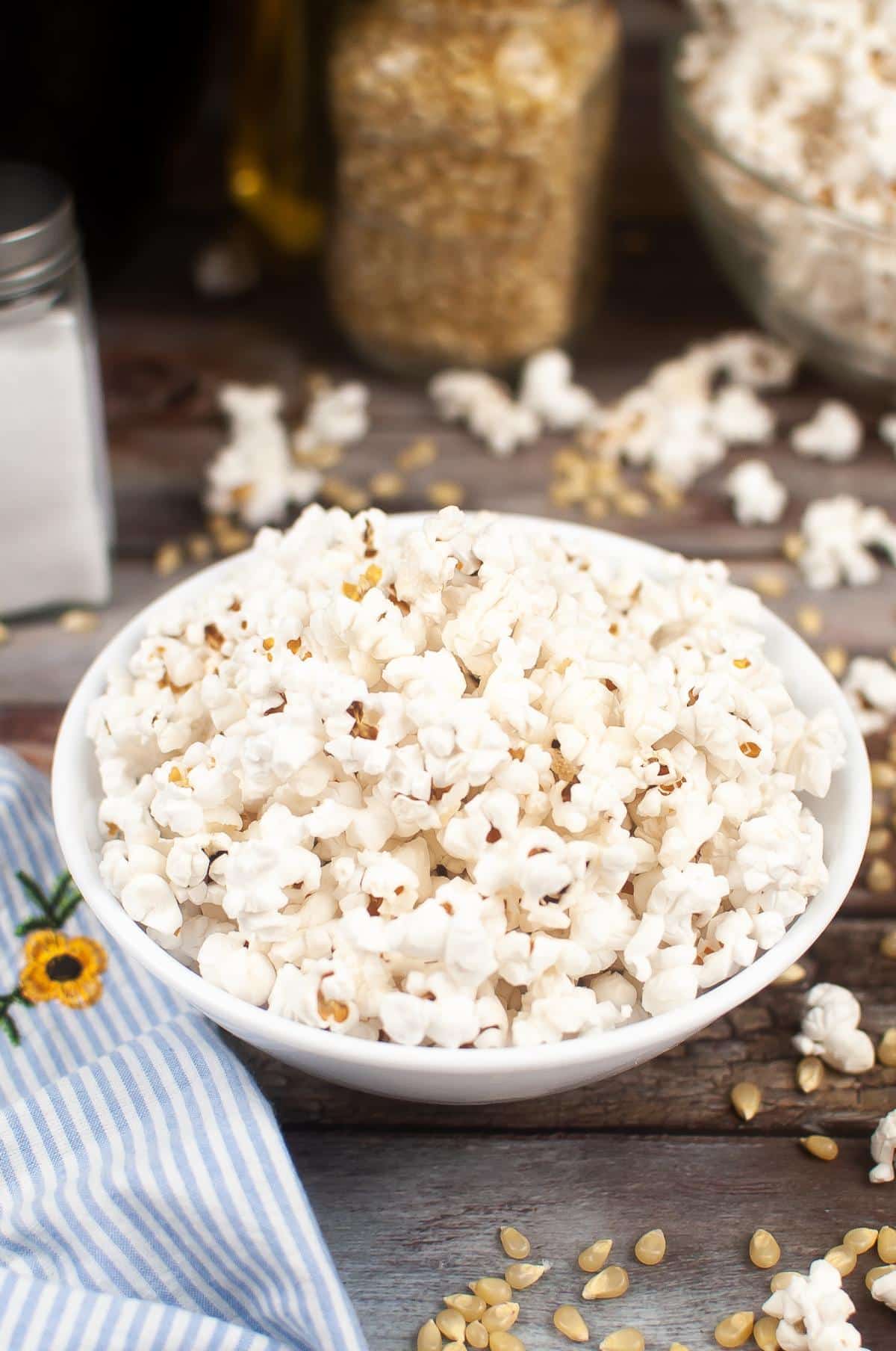 Bowl of air fryer popcorn on a wooden surface with kernels and a blue striped cloth nearby.
