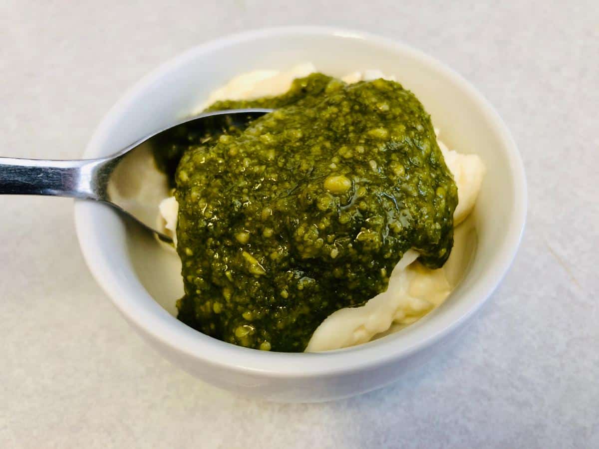 Mayonnaise and pesto sauce in a small white bowl with spoon in it.