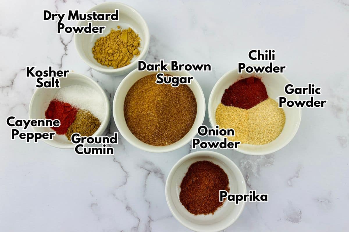 Bowls of spices and seasonings for pork dry rub on a marble countertop.