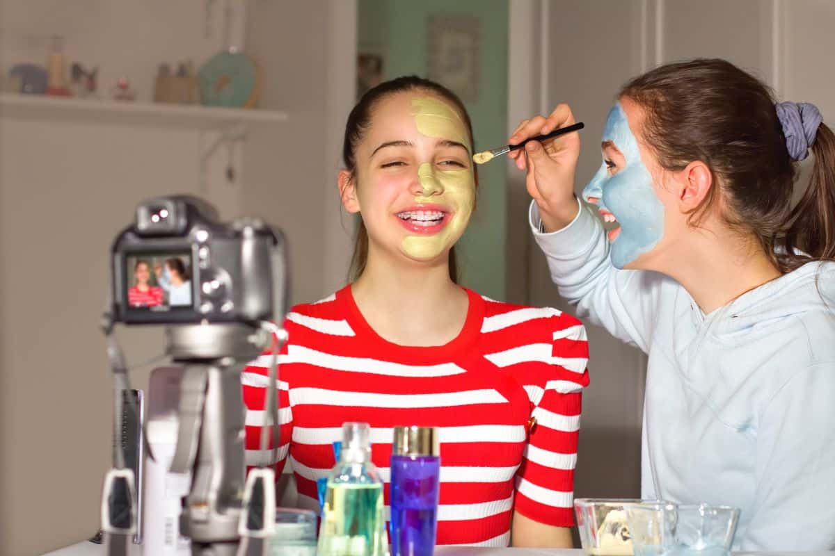 A girl is getting her face painted by her friend in front of the camera, a fun indoor activity for teens.