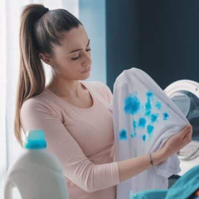 A woman is holding a blue shirt in front of a washing machine, trying to remove detergent stains.