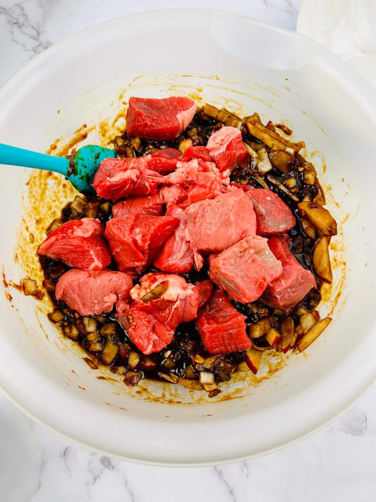 Raw beef is added into the marinade mixture in a bowl with a blue spatula.
