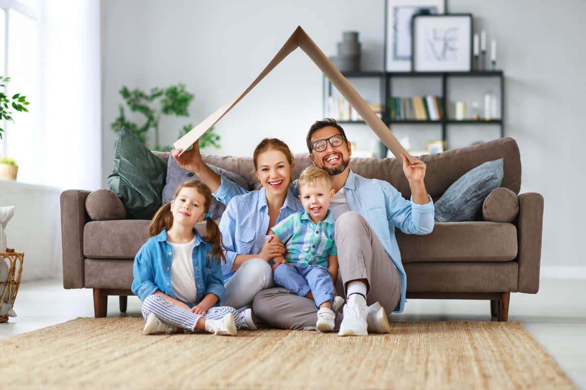A family sitting on the floor in their living room, with a cardboard house.